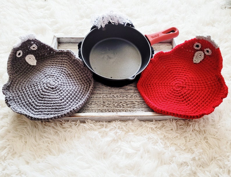 Pode incluir: Um conjunto de pegadores de croch&ecirc; em forma de galinhas, um cinzento e outro vermelho, com uma panela preta e uma pega vermelha. Os pegadores t&ecirc;m detalhes brancos e cinzentos. O conjunto est&aacute; numa bandeja de madeira.