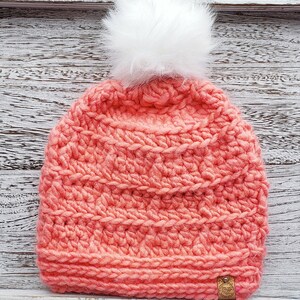 K&ouml;nnte beinhalten: Eine korallenrosa gestrickte M&uuml;tze mit einem wei&szlig;en Bommel oben. Die M&uuml;tze hat ein strukturiertes Stichmuster und ein kleines braunes Etikett an der Seite.