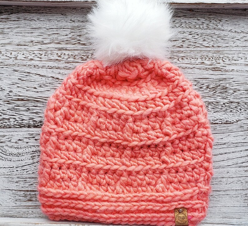 K&ouml;nnte beinhalten: Eine korallenrosa gestrickte M&uuml;tze mit einem wei&szlig;en Bommel oben. Die M&uuml;tze hat ein strukturiertes Stichmuster und ein kleines braunes Etikett an der Seite.