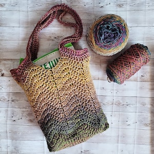 Op de afbeelding: Een gehaakte draagtas met een meerkleurig ombre-ontwerp in tinten bruin, groen en geel. De tas is gemaakt van een dik garen en heeft een brede, stevige band. De tas ligt op een houten oppervlak met twee bollen garen.
