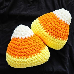 May include: Two crocheted candy corn pillows in yellow, orange, and white. The pillows are made with a fluffy yarn and have a three-dimensional look.