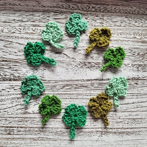 May include: A collection of nine crocheted shamrock shapes in various shades of green and brown, arranged in a circle on a whitewashed wooden surface.