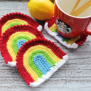 Puede incluir: Juego de cuatro posavasos de arcoíris de ganchillo hechos a mano. Cada posavasos presenta un vibrante diseño de arcoíris con arcos rojos, naranjas, amarillos, verdes y azules, bordeados de blanco y rojo. Una taza roja con el logo de Starbucks está encima.