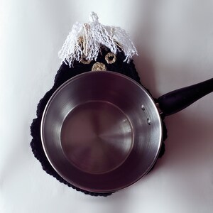 Pode incluir: Um pegador de panela preto e branco de croch&ecirc; com um design extravagante, apresentando um rosto com cabelo de franja branca e detalhes dourados. Uma panela de a&ccedil;o inoxid&aacute;vel com uma al&ccedil;a preta repousa sobre o pegador de panela.