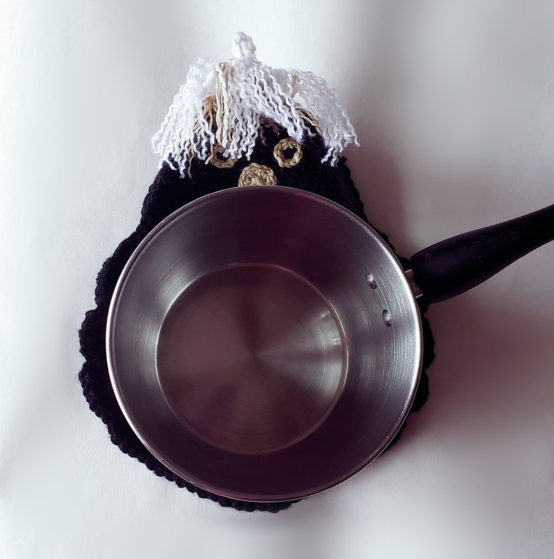 Pode incluir: Um pegador de panela preto e branco de croch&ecirc; com um design extravagante, apresentando um rosto com cabelo de franja branca e detalhes dourados. Uma panela de a&ccedil;o inoxid&aacute;vel com uma al&ccedil;a preta repousa sobre o pegador de panela.
