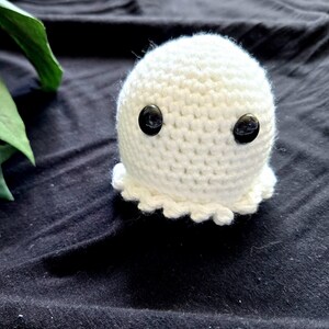 May include: A small, handmade white crochet ghost with two black button eyes. The ghost has a ruffled bottom edge and is set against a dark background. A few green leaves are visible in the background.
