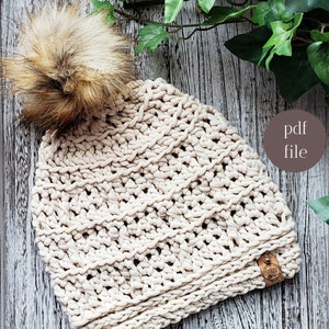 Puede incluir: Un gorro de punto beige con un pompón marrón esponjoso en la parte superior. El gorro tiene un patrón de punto texturizado y una etiqueta de cuero marrón con un logotipo. El gorro está fotografiado sobre una superficie de madera con hojas verdes.