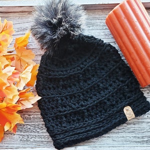 K&ouml;nnte beinhalten: Schwarze H&auml;kelm&uuml;tze mit einem flauschigen schwarzen Bommel oben. Die M&uuml;tze hat ein strukturiertes, geripptes Design und ein kleines braunes Leder-Etikett.