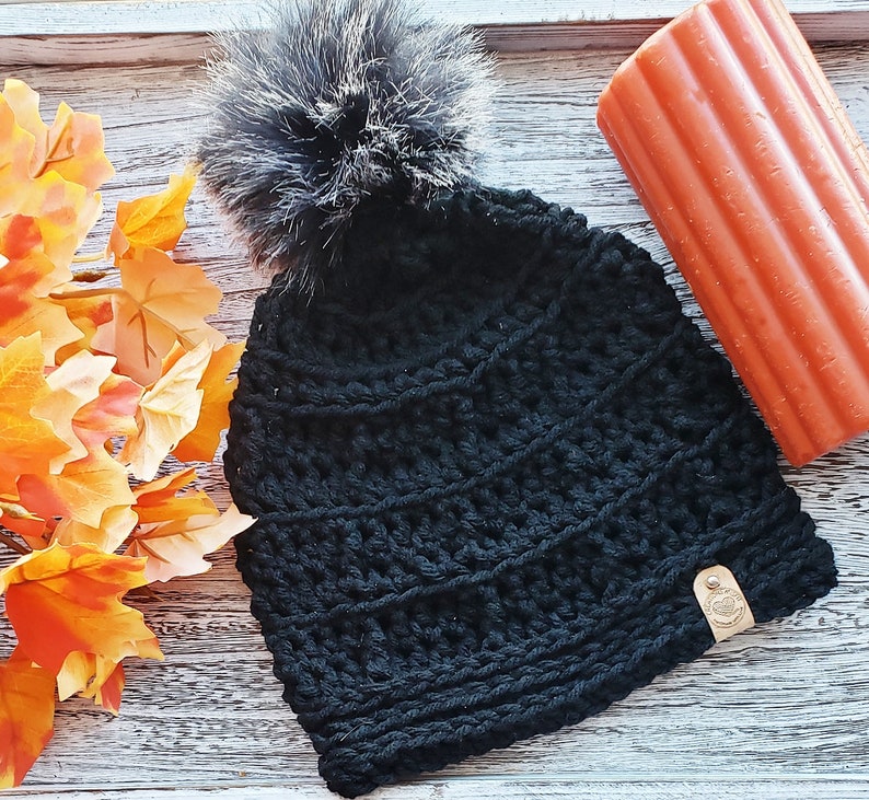 K&ouml;nnte beinhalten: Schwarze H&auml;kelm&uuml;tze mit einem flauschigen schwarzen Bommel oben. Die M&uuml;tze hat ein strukturiertes, geripptes Design und ein kleines braunes Leder-Etikett.