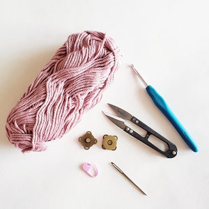 Puede incluir: Una madeja de hilo rosa, un par de tijeras peque&ntilde;as, dos cierres magn&eacute;ticos dorados, un marcador de punto rosa, un gancho de ganchillo y una aguja sobre una superficie blanca.