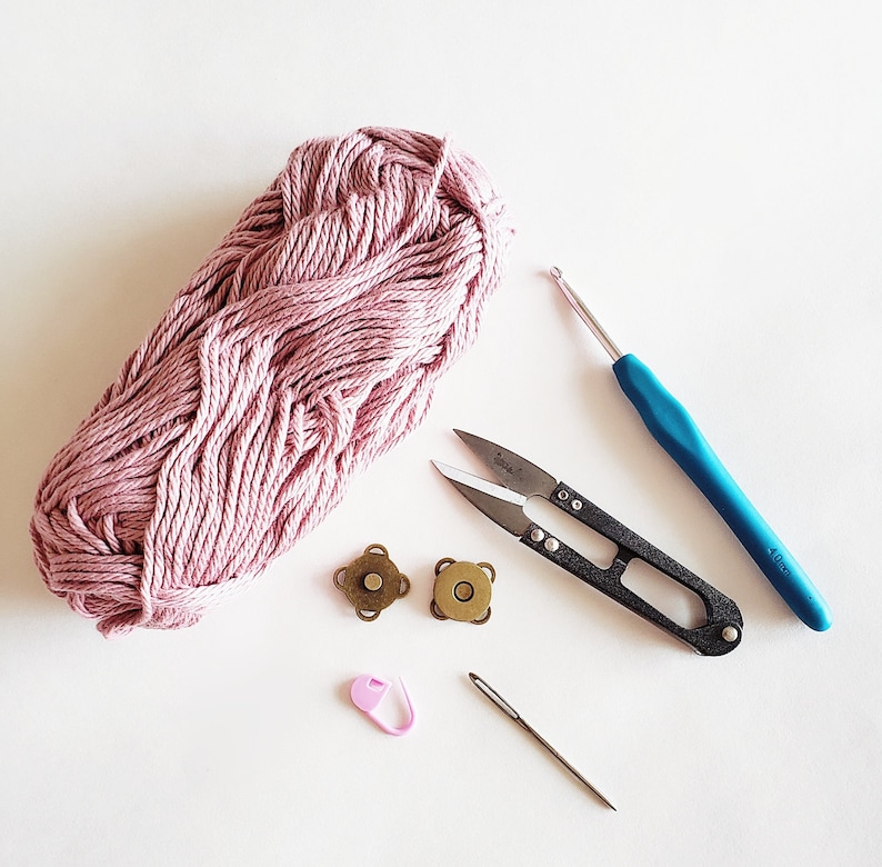 Puede incluir: Una madeja de hilo rosa, un par de tijeras peque&ntilde;as, dos cierres magn&eacute;ticos dorados, un marcador de punto rosa, un gancho de ganchillo y una aguja sobre una superficie blanca.