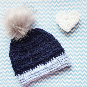 K&ouml;nnte beinhalten: Eine marineblaue H&auml;kelm&uuml;tze mit einem hellblauen Rand und einem flauschigen wei&szlig;en Bommel. Im Hintergrund befindet sich ein wei&szlig;es H&auml;kelherz.