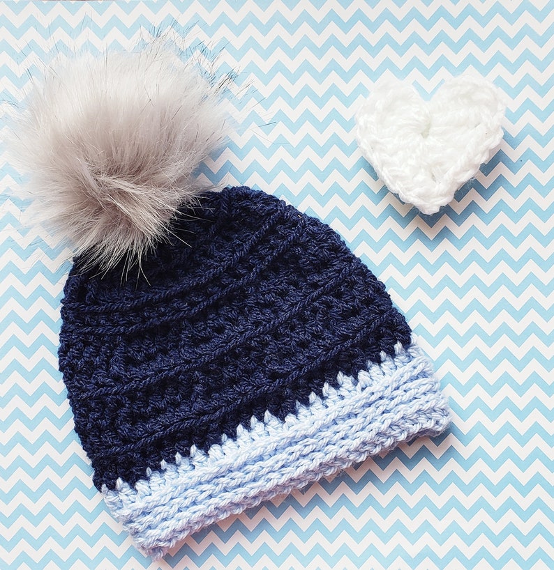 K&ouml;nnte beinhalten: Eine marineblaue H&auml;kelm&uuml;tze mit einem hellblauen Rand und einem flauschigen wei&szlig;en Bommel. Im Hintergrund befindet sich ein wei&szlig;es H&auml;kelherz.