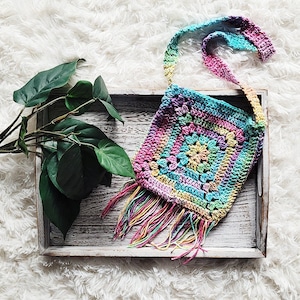 May include: A colorful crocheted bag with a fringe. The bag is made with a variety of colors, including pink, blue, green, and yellow. The bag has a long strap and is sitting on a wooden surface.