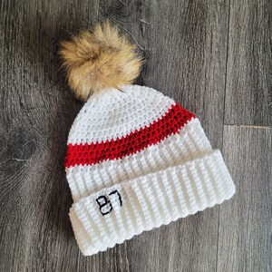 May include: White crocheted beanie with a red stripe and a brown pom pom on top. The beanie has the number "87" embroidered on the front.