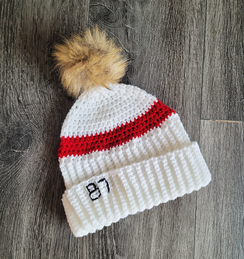 May include: White crocheted beanie with a red stripe and a brown pom pom on top. The beanie has the number "87" embroidered on the front.