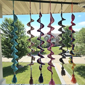 May include: Six hanging crochet spiral ornaments suspended from a black rod. Each spiral features a unique color scheme, including solid turquoise, white and brown, red white and blue, pink and white, grey, and a multi-colored rainbow pattern.