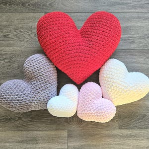 May include: A collection of handmade crochet heart-shaped pillows in various sizes and colors. The largest pillow is red, with others in lavender, white, and pink. The pillows are arranged on a wooden surface.