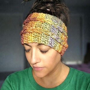 Puede incluir: Una diadema de ganchillo hecha a mano, multicolor, en tonos amarillos, naranjas, verdes y morados. La diadema se lleva en la cabeza y tiene un aspecto texturizado y tejido. La persona que lleva la diadema lleva una camiseta verde.