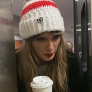 May include: A woman wearing a white and red knitted beanie with the number "87" embroidered on it, holds a white coffee cup and a pound note.