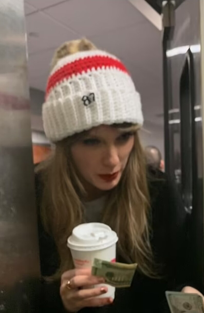 May include: A woman wearing a white and red knitted beanie with the number "87" embroidered on it, holds a white coffee cup and a pound note.