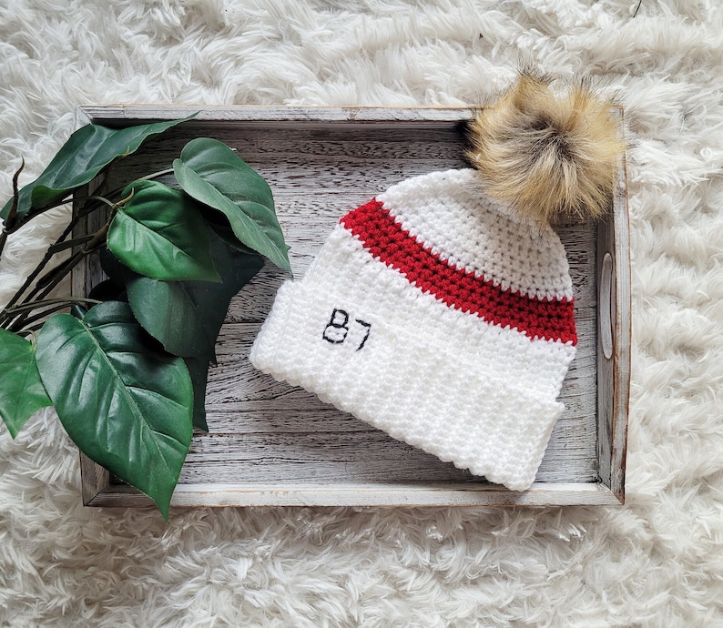 May include: White crocheted hat with a red stripe and the number 87 embroidered on the front. The hat has a fluffy pom pom on top.