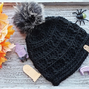 K&ouml;nnte beinhalten: Schwarze H&auml;kelm&uuml;tze mit einem flauschigen schwarzen Bommel. Die M&uuml;tze hat ein strukturiertes Stichmuster und ein kleines Leder-Etikett mit einem Herz-Design.
