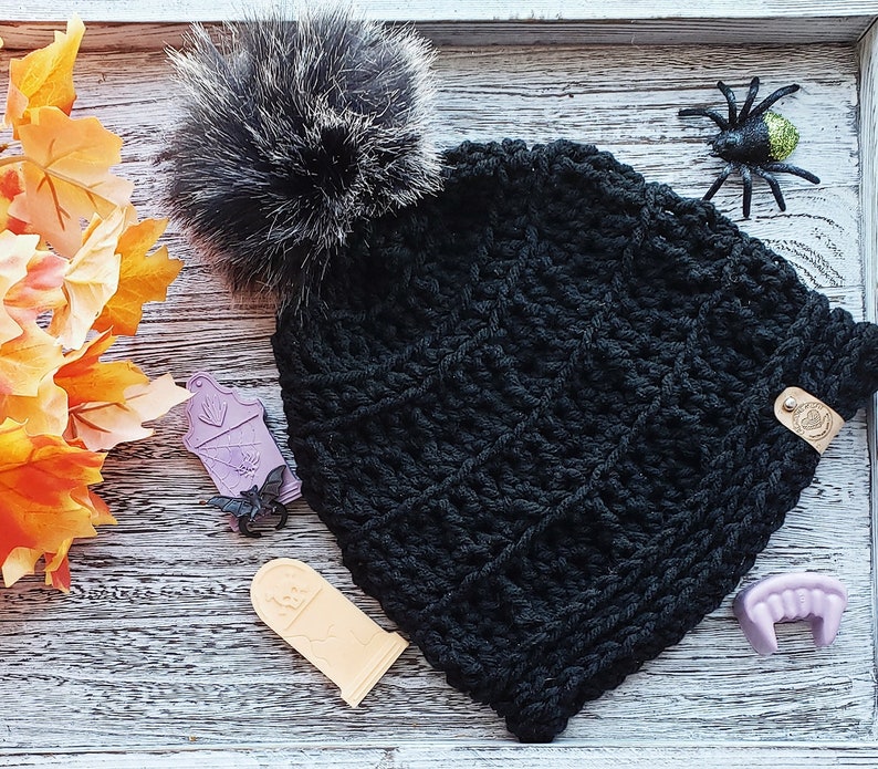 K&ouml;nnte beinhalten: Schwarze H&auml;kelm&uuml;tze mit einem flauschigen schwarzen Bommel. Die M&uuml;tze hat ein strukturiertes Stichmuster und ein kleines Leder-Etikett mit einem Herz-Design.