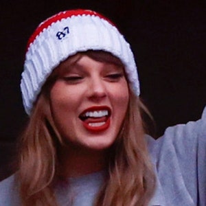 May include: A woman wearing a white and red knitted beanie with the number "87" embroidered on it. She is smiling and wearing a grey sweatshirt.