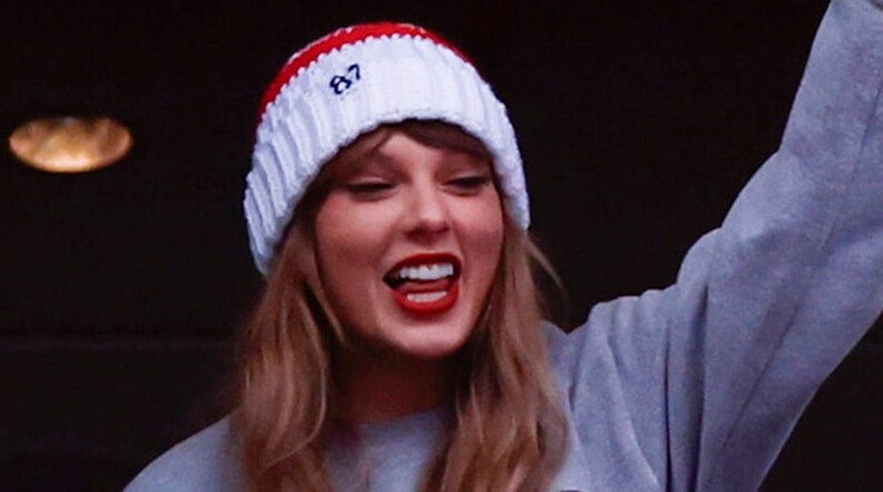 May include: A woman wearing a white and red knitted beanie with the number "87" embroidered on it. She is smiling and wearing a grey sweatshirt.