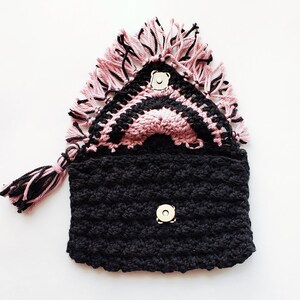 Puede incluir: Un bolso de crochet negro con una solapa de crochet rosa y negro. El bolso tiene un detalle de flecos y dos cierres a presi&oacute;n.