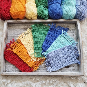 Puede incluir: Un conjunto de siete posavasos de ganchillo en un arcoíris de colores, incluyendo rojo, naranja, amarillo, verde, azul, azul claro y morado. Cada posavasos tiene un borde de flecos y está hecho con un patrón de punto diferente.