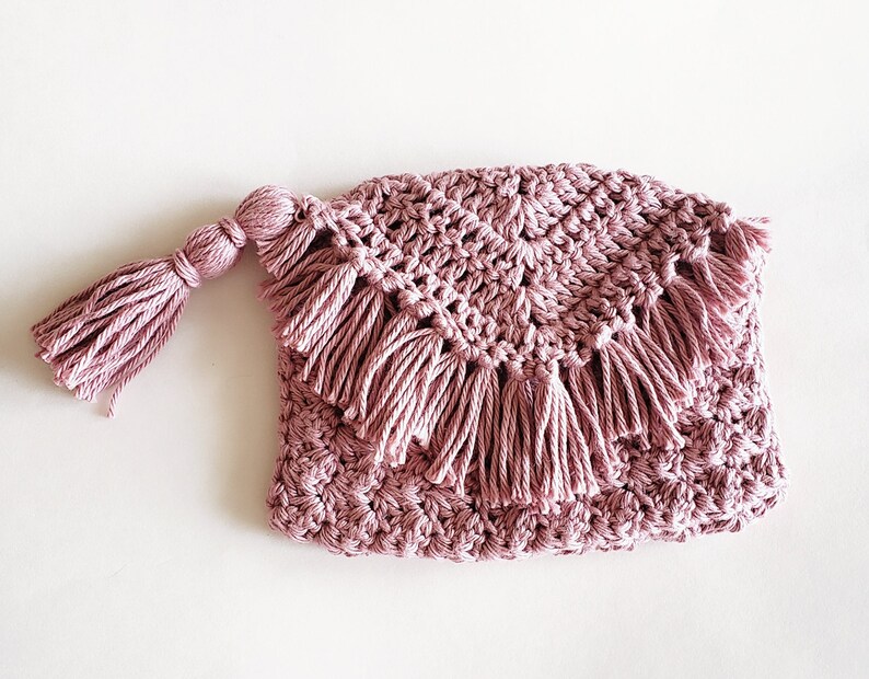 Puede incluir: Una bolsa de crochet rosa con un borla. La bolsa tiene un borde festoneado y est&aacute; decorada con flecos.