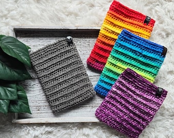 May include: Five crocheted striped phone cases in a rainbow color scheme. The cases are arranged on a wooden tray with a gray crocheted case in the background.