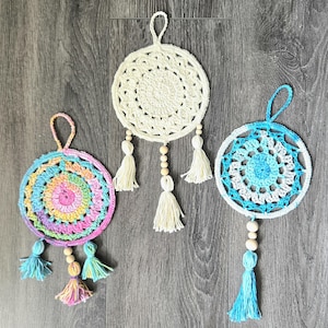 May include: Three handmade crochet dream catchers. One is cream-colored, another is a rainbow of colors, and the third is blue and white. Each has a loop for hanging, wooden beads, and tassels. The dream catchers are displayed on a wooden surface.