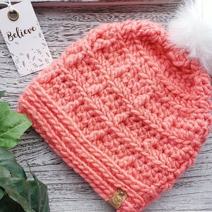 K&ouml;nnte beinhalten: Eine korallenrosa gestrickte M&uuml;tze mit einem wei&szlig;en Bommel oben. Die M&uuml;tze hat ein strukturiertes Stichmuster und ein kleines braunes Leder-Etikett, das an der Seite befestigt ist. Das Etikett lautet "Believe".