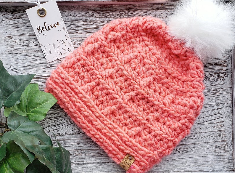 K&ouml;nnte beinhalten: Eine korallenrosa gestrickte M&uuml;tze mit einem wei&szlig;en Bommel oben. Die M&uuml;tze hat ein strukturiertes Stichmuster und ein kleines braunes Leder-Etikett, das an der Seite befestigt ist. Das Etikett lautet "Believe".