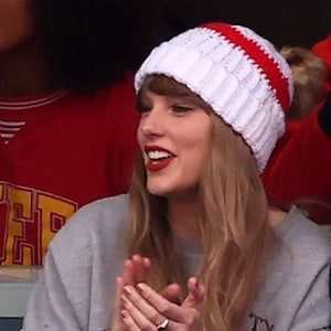 May include: A woman wearing a white and red knitted beanie with a pom pom on top. She is wearing a grey sweatshirt and has long blonde hair.