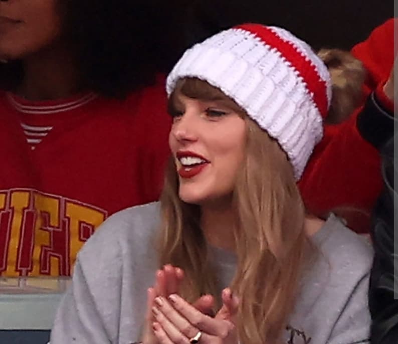 May include: A woman wearing a white and red knitted beanie with a pom pom on top. She is wearing a grey sweatshirt and has long blonde hair.