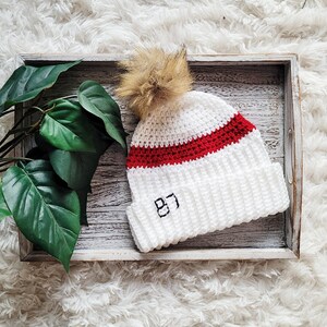 May include: White crocheted beanie with a red stripe and a brown pom pom. The beanie has the number "87" embroidered on the front.