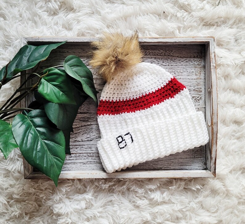 May include: White crocheted beanie with a red stripe and a brown pom pom. The beanie has the number "87" embroidered on the front.
