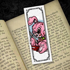 May include: A pink bunny bookmark with a syringe, blood, and a stitched design. The bookmark has a black border with a white and blue background. The bunny has a stitched design and is holding a syringe.