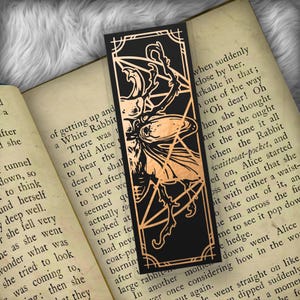 May include: Black bookmark with a copper-colored illustration of a figure with a bow and arrow, set within a geometric frame. The bookmark is rectangular and rests on the open pages of a book.