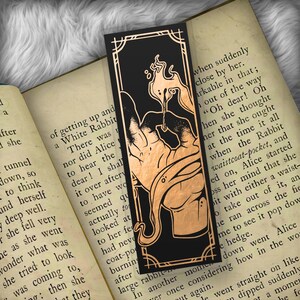 May include: Black bookmark with a copper-colored illustration of a creature and a decorative border. The bookmark is resting on an open book with text. The illustration is a stylized design.