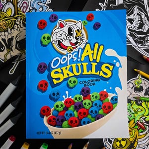 May include: A blue coloring book titled "Oops! All Skulls" features a cartoon cat mascot and colorful skull-shaped cereal. The cover art includes red, purple, green, and blue skulls. The book is surrounded by art supplies and black illustrations.