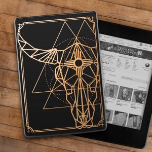 May include: Black e-reader cover with a gold geometric bull skull design. The cover is rectangular with a gold border. The e-reader screen displays the Etsy shop "SkullsAndSucculents". The background is a wooden surface.