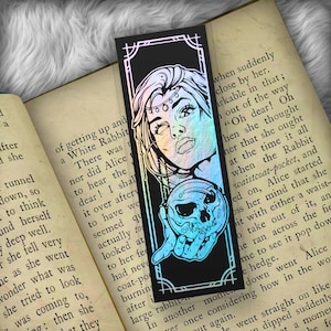 May include: A black bookmark with a holographic finish features a woman with a skull in her hand. The woman has a crown of flowers and a jeweled necklace. The text on the bookmark reads "Oh dear! Oh when she thought that she ought at the time it all when the Rabbit waistcoat-pocket, and ed on, Alice started her mind that she bit with either a waist- to take out of it, an he ran across the fie me to see it pop dow e hedge. down went Alice how in the wor ver once considering t again."