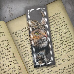 May include: A bookmark featuring a detailed illustration of a cat wearing a Viking helmet and ornate jewelry. The cat has orange and brown fur with green eyes, set against a gray background with a white border. The bookmark is resting on the pages of a book.