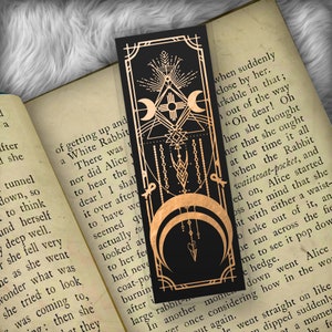 May include: A black bookmark with a gold design featuring a crescent moon, a sun, and a geometric pattern. The bookmark has the text "when suddenly close by her. arkable in that; out of the way "Oh dear! Oh hen she thought her that she ought at the time it all when the Rabbit waistcoat-pocket, and ed on, Alice started oss her mind that she it with either a waist- o take out of it, an he ran across the fie In another mome me to see it pop dow down went Alice e hedge. ver once considering how in the wor t again. le went straight on like dipped suddenly ot a momen he fo"