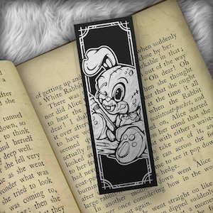 May include: Black and white bookmark featuring a cartoon rabbit with a syringe, set against a decorative frame. The bookmark is placed on an open book with text visible.